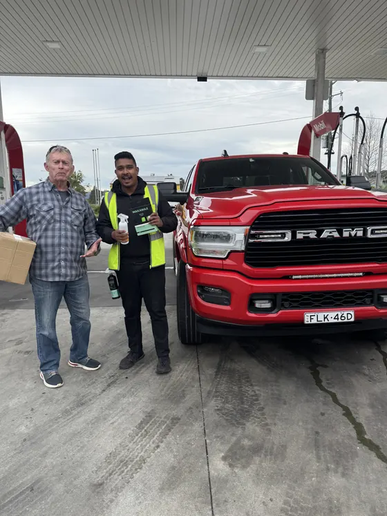 A WakaWax customer and seller with a box of WakaWax beside a red pickup truck at a service station