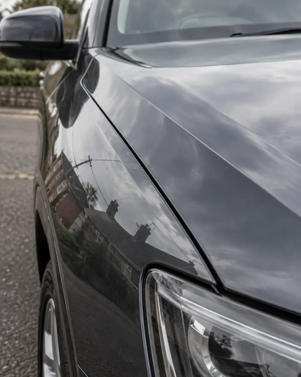 The glossy front wing of a dark car reflecting a cloudy sky